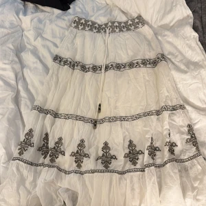 Vit långkjol med broderi från Zara - Supersnygg vit långkjol från Zara med svart broderat mönster och dekorativa band. Kjolen har volanger och en justerbar snörning i midjan. Tillverkad i lätt bomull som ger en flowy och chill vibe. Perfekt för dig som gillar boho-stil och detaljer.