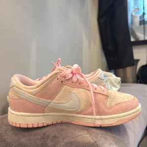 Nike Dunk Low i rosa och beige mocka - Nike Dunk Low sneakers i en snygg kombo av ljusrosa och beige mocka med vita detaljer och rosa snören. Klassisk låg siluett med rund tå och platt sula. Perfekta för dig som vill sticka ut med en soft pastell-look.