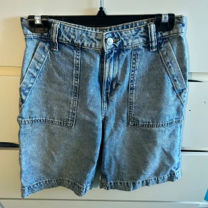 Blå jeansshorts från DIVIDED - Snygga blå jeansshorts från DIVIDED med loose passform och hög midja. Klassisk femficksdesign med stora fickor fram och bak, samt bälteshällor och dragkedja. Perfekt tvättad denim-look som ger en cool vibe till din outfit.