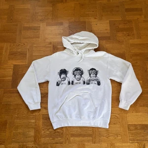Vit hoodie med aptryck från Gildan - Vit hoodie från Gildan med tryck av tre apor och texten 'Don't see, don't hear, don't speak' på bröstet. Klassisk känguruficka framtill, huva med snörning och långa ärmar. Perfekt för dig som gillar streetstyle och coola prints.