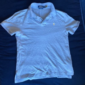 Ljusblå Polo Ralph Lauren piké storlek 160 - Snygg ljusblå t-shirt från Polo Ralph Lauren med klassisk krage och korta ärmar. Slim fit-modell med gul broderad logga på bröstet. Tillverkad i mjuk bomull som är skön mot huden. Perfekt för dig som gillar stilren och enkel design.