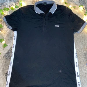 Svart BOSS t-shirt med grå krage - Svart slim fit t-shirt från BOSS med stretch, gråmelerad krage och ärmslut. Snygga vita band med BOSS-logga längs sidorna. Klassisk pikékrage och korta ärmar, perfekt för en clean och sportig look. Fläcken ni ser kommer jag ta bort om ni köper 