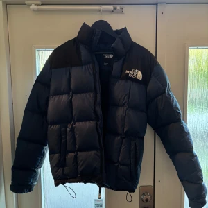  The North Face  - Säljer en blå och svart dunjacka från The North Face i storlek XS. Jackan har en klassisk pufferstil med hög krage, dragkedja och broderad logga på bröstet. Perfekt för kalla dagar.