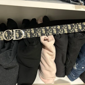 Svart och beige Dior bälte med monogram - Säljer ett snyggt Dior bälte med klassiskt monogrammönster i svart och beige. Bältet har en silverfärgad metallspänne med Dior-logga och insida i svart skinn. Perfekt accessoar för att lyfta din outfit med lyxig känsla.