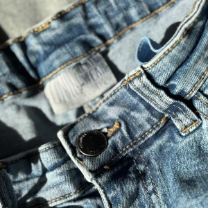 Blå bootcut jeans från United Wear - Snygga ljusblå bootcut jeans från United Wear med klassisk femficksdesign och hög midja. Jeansen har en lätt tvättad look, raka ben som blir vidare nedtill och är tillverkade i mjukt denimtyg. Perfekta för en avslappnad och trendig stil.