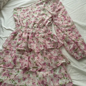 Blommig one shoulder klänning - Superfin kort blommig klänning från Meridian i storlek 38. One shoulder-modell med lång ärm, volanger och smock i midjan. Ljusrosa och grönt blommönster på vit botten, perfekt för vår och sommar. Luftigt material och snygg passform.