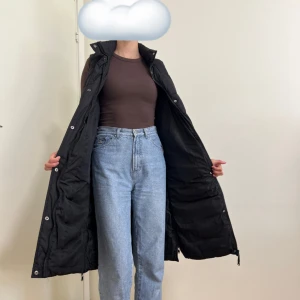 Lång svart dunväst med avtagbar huva - Lång svart dunväst med huva och hög krage. Västen är quiltad och har dragkedja hela vägen samt tryckknappar. Jätteskön, som ny då jag endast använt den en höst. Passar S och M. Pris kan diskuteras 💖