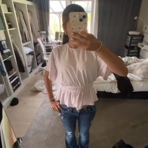 Ljusrosa blus med volang - Supersöt ljusrosa blus med korta, vida ärmar och smock i midjan som ger en snygg volang nertill. Perfekt att styla med jeans eller kjol för en fräsch look. Tillverkad i mjuk bomull som känns skön mot huden.