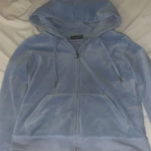 Ljusblå velour hoodie Juicy Couture - Supermjuk ljusblå hoodie från Juicy Couture i velour med dragkedja och huva. Klassisk broderad JC-logga på bröstet, snörning i huvan och känguruficka framtill. Perfekt för en chill och trendig look. Lite trasig längst upp i dragkedjan men det går att fixa. 