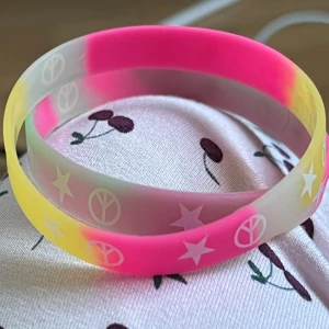 Färgglada silikonarmband med stjärnor - Två coola armband i silikon med färgskiftningar i rosa, gul och vit. Armbanden har tryckta vita stjärnor och peace-symboler runt om. Perfekt accessoar för att ge din outfit en lekfull och unik touch.