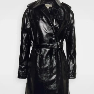 Svart lack trenchcoat med bälte - Säljer en svart glansig trenchcoat i lack-look med bälte i midjan och klassisk krage. Jackan har dubbelknäppning, axelklaffar och spännen vid ärmsluten för extra edge. Perfekt för dig som vill sticka ut med en cool och tidlös stil.