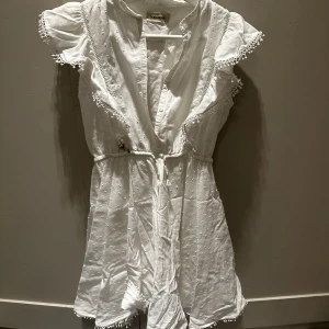 Klänning från Zadig&voltaire - Supersöt vit kortklänning från Jaba Yard med broderade detaljer och spetskant på ärmarna. Klänningen har knappar framtill, snörning i midjan och volangärmar. Perfekt för sommarens alla tillfällen och ger en fräsch och romantisk vibe.