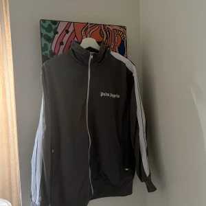 Palm Angels grå vindjacka med vita ränder - Palm Angels vindjacka i grått med vita ränder längs ärmarna och dragkedja framtill. Jackan har hög krage och logga på bröstet. Perfekt för dig som gillar streetwear och vill sticka ut med en clean look.