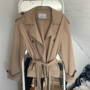 Beige trenchcoat från Stradivarius - Säljer en snygg beige trenchcoat från Stradivarius med klassisk krage, dubbelknäppning och bälte i midjan. Jackan har axelklaffar, knappar vid ärmslut och en kortare, figurnära passform. Perfekt för vår och höst!