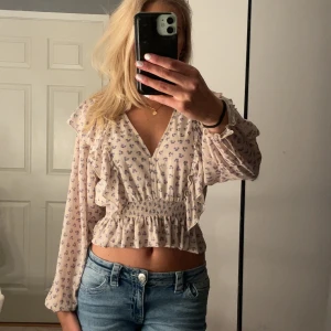 Blommig volangblus med v-ringning - Supersöt blommig blus i ljusbeige med små lila blommor, v-ringning och volangdetaljer längs bröstet.🥹🩷