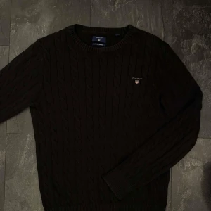 Mörkbrun kabelstickad tröja GANT - Snygg mörkbrun kabelstickad tröja från GANT med rund halsringning och diskret logga broderad på bröstet. Tröjan har långa ärmar och ribbade muddar, perfekt för lager-på-lager under kyliga dagar.