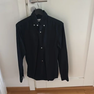 Svart skjorta från John Henric - Stilren svart skjorta från John Henric med klassisk krage och vita knappar. Skjortan är långärmad och har en snygg, tidlös design som passar perfekt till jeans eller kostymbyxor. Tillverkad i mjuk bomull för en skön känsla hela dagen.