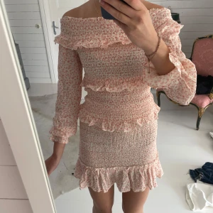 Blommig offshoulder klänning Bik Bok - Supersöt kort offshoulder klänning från Bik Bok i vitt och ljusrosa med blommigt mönster. Smockad passform, volangdetaljer och långa ärmar. Perfekt för sommarens alla tillfällen. Säljer då den inte kommer till användning längre!