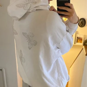 Vit hoodie med strassfjärilar - Superfin vit hoodie med dragkedja och huva, dekorerad med glittriga strassfjärilar på både ärmar, rygg och huva. Tröjan har kängurufickor och en relaxed passform, perfekt för dig som gillar en söt och trendig stil.