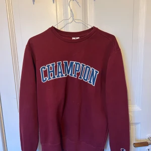 Mörkröd sweatshirt från Champion - Säljer en mörkröd sweatshirt från Champion i storlek small. Tröjan har klassisk rund hals, långa ärmar och stor CHAMPION-logga i vitt och blått framtill. Mjuk insida och ribbade muddar vid ärmslut och nederkant. Perfekt för chill dagar.