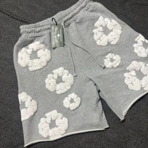 Grått set från Denim Tears med vita mönster - Grå t-shirt från Denim Tears med coola vita applikationer i form av molnliknande cirklar över hela tröjan. T-shirten har rund hals och korta ärmar, tillverkad i mjuk bomull för skön känsla. Perfekt för dig som gillar streetwear och unika prints.