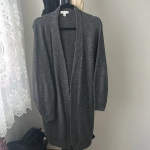 Mörkgrå stickad kofta från H&M - Säljer en mörkgrå, lång och mysig stickad kofta från H&M i storlek S. Koftan har en öppen front utan knappar och långa ärmar, perfekt att slänga på sig när det är lite kyligt. Enkel och stilren design som funkar till många outfits.