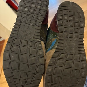 Valentino Garavani camo sneakers - Snygga Valentino Garavani sneakers med camouflagemönster i blått, grönt och beige. Skorna har detaljer i mocka och skinn, samt coola guld- och vinröda partier vid hälen. Klassisk snörning och platt sula. Perfekta för dig som vill sticka ut med stil.