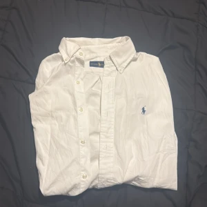 Vit Ralph Lauren - Klassisk vit skjorta från Ralph Lauren med blå broderad logga på bröstet. Skjortan har knappar hela vägen, button down-krage och är tillverkad i bomull. Perfekt för en clean och stilren look.
