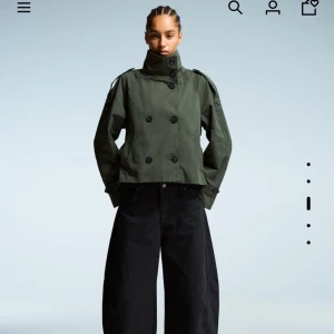 Olivgrön kort trenchcoat från Bershka - Säljer en olivgrön kort trenchcoat från Bershka med dubbelknäppning, axelklaffar och bälte i midjan. Jackan har en loose passform och är tillverkad i bomullsmix. Perfekt för dig som vill ha en trendig och cool look. Den är helt slutsåld och oanvänd därav priset! 