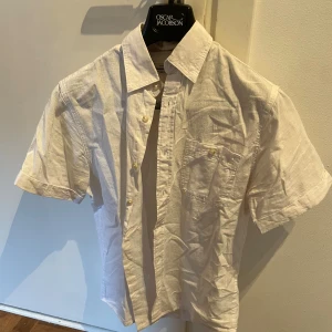 Vit kortärmad linneskjorta Dressman - Vit skjorta i linne från Dressman med korta ärmar, klassisk krage och bröstficka. Perfekt för varma dagar och har snygga knappar framtill. Enkel och stilren design som passar till mycket.