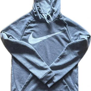 Grå Nike hoodie med stor logga - Snygg grå hoodie från Nike med stor vit Swoosh-logga på bröstet. Tröjan har huva med snörning och är tillverkad i mjukt Dri-FIT-material som andas. Perfekt för dig som gillar sportig och avslappnad stil.