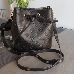 Svart bucket axelväska - Snygg svart bucket axelväska med klassiskt monogram. Väskan har justerbar axelrem med guldfärgade detaljer och stängs med snörning upptill. Perfekt storlek för vardagens essentials och ger en lyxig touch till din outfit.