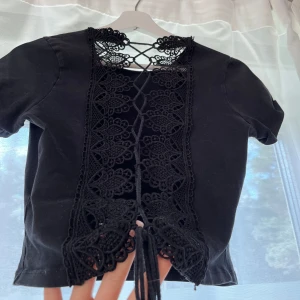 Svart topp med spets och snörning - Svart croppad topp med korta ärmar och unik rygg i spets med snygg snörning. Toppen har en cool öppen rygg med dekorativa spetsdetaljer och knyt längst ner. Perfekt för dig som vill sticka ut med en edgy och trendig look.