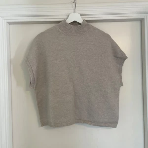 Beige stickad väst med hög krage - En stilren beige stickad väst med hög ribbad krage och breda ärmöppningar. Västen har en rak och loose passform, perfekt att slänga över en skjorta eller t-shirt. Ribbstickade detaljer vid krage, ärmöppningar och nederkant ger en snygg touch.