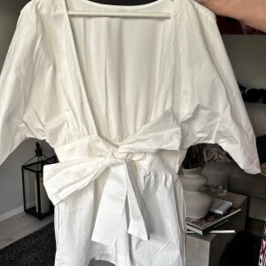 Vit blus med knyt - Snygg vit blus med omlott-design och bred knytning i midjan. Blusen har korta ärmar och ett luftigt, lätt material som ger en fräsch och modern vibe. Perfekt för dig som gillar stilrena och enkla plagg med en twist. Passar dig som vill ha en feminin look med cool edge.