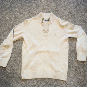 Beige stickad half zip-tröja Gant - Snygg beige stickad tröja från Gant med half zip-dragkedja och ribbade muddar. Klassisk design med liten broderad logga på bröstet och hög krage. Perfekt för lager-på-lager och chillare dagar. Nypris ca 800 kr