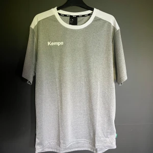 Grå sportig t-shirt från Kempa XL - Sportig t-shirt från Kempa i mörkgrå melange med vita detaljer på axlar och krage. Klassisk rund halsringning och korta ärmar. Perfekt för träning eller chill dagar. Tillverkad i lätt polyester som andas och känns skön mot huden. Tröjan är oanvänd då jag beställde fel storlek.