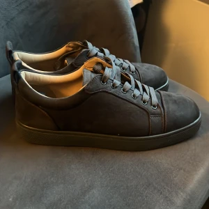 Louboutin skor mörk grå - Säljer ett par svarta sneakers från Christian Louboutin med klassisk röd sula och diskret logga undertill. Skorna har snörning, rund tå och är tillverkade i mocka med en stilren design. Perfekta för dig som vill ha något exklusivt och snyggt till din outfit.