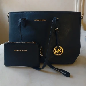 Mörkblå axelväska Michael Kors - Aldrig använd. Snygg mörkblå axelväska från Michael Kors i strukturerat skinn med guldfärgade detaljer och logotyp Michael kors på alla dragkedjor. Väskan har dragkedja upptill, flera innerfack och medföljande matchande plånbok. Perfekt storlek för allt du behöver och stilren design.