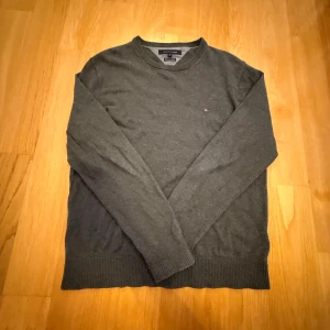 Mörkgrå tröja Tommy Hilfiger M - Mörkgrå långärmad tröja från Tommy Hilfiger i mjuk pima bomull och kashmir. Klassisk rund halsringning, diskret logga broderad på bröstet och ribbade muddar. Perfekt för dig som gillar stilren och enkel design.