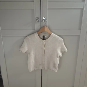 Beige kortärmad kofta från H&M Divided - Kortärmad beige kofta från H&M Divided i storlek S. Tröjan har rund hals, knappar framtill och är stickad i mjukt material. Perfekt för lager-på-lager och har en clean, enkel look som funkar till många stilar.