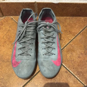 Gråa Nike fotbollsskor med rosa swoosh - Nike Air Zoom mercurial Vapor 16 Pro  - Säljer ett par gråa Nike fotbollsskor med en stor rosa swoosh på sidan. Sulan har gröna dobbar för bra grepp på planen. Perfekta för dig som vill sticka ut på matchen! Stolek 41.