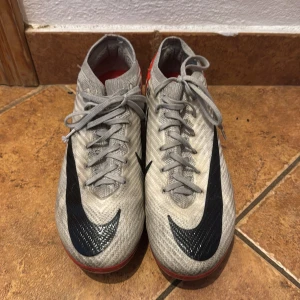 Nike fotbollsskor i grått och rött - Nike Court Zoom Vapor 15 - Snygga Nike fotbollsskor med grå ovandel, stora svarta swoosh-loggor och röda detaljer på sidorna och sulan.