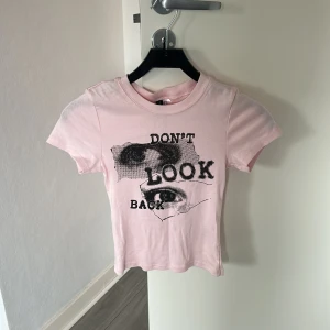 Rosa t-shirt med tryck 'Don't Look Back' - Snygg ljusrosa t-shirt med coolt svart tryck och texten 'Don't Look Back' på framsidan. Modellen är croppad och har rund hals samt korta ärmar. Perfekt för dig som gillar statement-plagg och streetstyle.