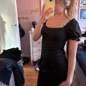 Svart kort klänning från NA-KD - Säljer en svart kort klänning från NA-KD i storlek 36. Klänningen har puffiga korta ärmar, fyrkantig urringning och spetsdetaljer på axelbanden. Sitter snyggt och har en slim passform som framhäver figuren. Perfekt för dig som gillar stilrena och trendiga plagg. Använt en gång 