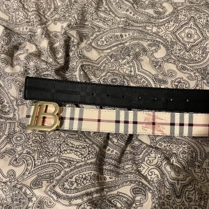 Burberry rutigt bälte med guldspänne - Snyggt Burberry bälte med klassiskt rutigt mönster i beige, svart och rött. Bältet har ett stilrent guldspänne med Burberrys ikoniska B-logga. Materialet är skinn och designen är både modern och tidlös, perfekt för att lyfta din outfit.