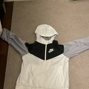 Nike vindjacka med huva, vit och grå - Nike vindjacka i vit och ljusgrå med svart panel och logga på bröstet. Jackan har huva, hel dragkedja och två sidofickor med dragkedja. Insidan är fodrad med mesh för extra ventilation. Perfekt för dig som gillar sportig stil.