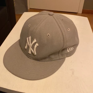 Grå New Era NY keps 59FIFTY - Säljer en stilren grå New Era 59FIFTY keps med New York Yankees-logga broderad framtill i vitt. Kepsen har klassisk form, böjd skärm och broderade ventilationshål. Tillverkad i 100% polyester och passar perfekt för dig som gillar sportig streetstyle.
