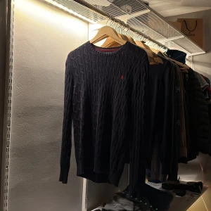 Mörkblå stickad tröja Polo Ralph Lauren - Säljer en mörkblå stickad tröja från Polo Ralph Lauren med klassiskt kabelstickat mönster och röd broderad logga på bröstet. Jag fick den i julklapp för över 1 år sedan och har tänkt att jag ska växa i den. Men nu har det gått så pass länge och den är fortfarande gigantisk på mig. Tröjan är köpt i USA och passar nog bäst om du är runt 180 cm lång!