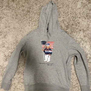 Grå hoodie Polo Ralph Lauren Team USA - Snygg grå hoodie från Polo Ralph Lauren med Team USA-tryck och ikoniska Polo Bear på bröstet. Tröjan har huva med snörning, lång ärm och patch på ena ärmen. Perfekt för dig som gillar sportig och avslappnad stil.